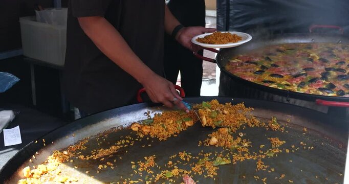 Street food vendor serves fresh seafood paella from large traditional metal pan into white plate. Chef hands use metal spatula to scoop yellow rice with mussels shrimps and vegetables during outdoor