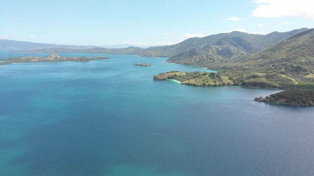 Aerial panorama of Pulau Rinca Komodo in Flores, Indonesia, featuring lush green hills surrounded by deep blue tropical waters under bright clouds and clear daylight skies