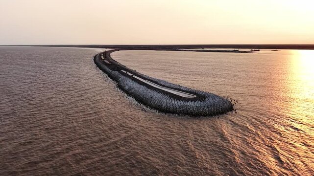 Slow panning aerial shot of new port breakwater construction extending into calm sea at golden sunset with warm reflections on water