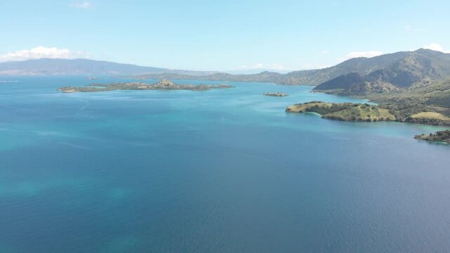 Aerial panorama of Pulau Rinca Komodo in Flores, Indonesia, featuring lush green hills surrounded by deep blue tropical waters under bright clouds and clear daylight skies