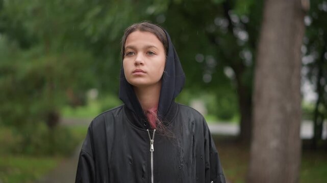 Young woman in soaked black hoodie pausing on treelined boardwalk, droplets sliding from hood while eyes close in calm resolve, student commuter vibe with cinematic shallow depth and textured wet