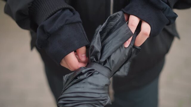 Hands untangling black umbrella after rain, closeup of woman manipulating compact canopy, water beads sliding off fabric, finger movements, zippered jacket, gusty street scene, practical urban moment