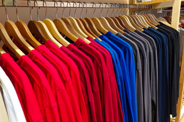 Fototapeta premium Neat rows of colorful shirts hang on wooden hangers. Bright reds, blues, and grays line a wooden clothing rack.