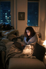 Fototapeta premium Woman working on a laptop under a blanket in a cozy apartment at night.