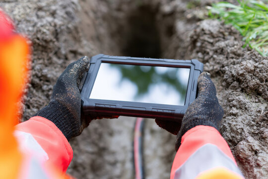 Rugged drain camera monitor held by gloved hands over muddy trench with visible pipe and green grass nearby