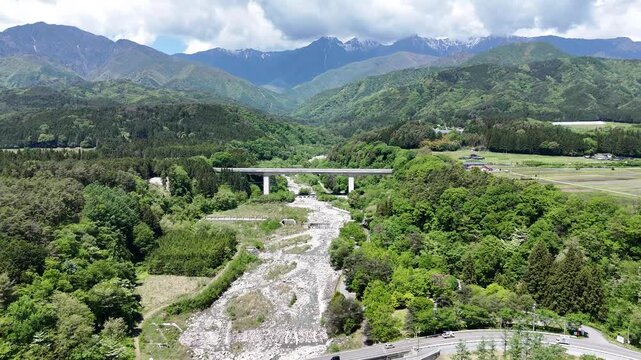 Green mountains frame a river's rushing path. Concrete bridges span the waterway with grace. Vast forests stretch beneath a partly cloudy sky. No people are visible here