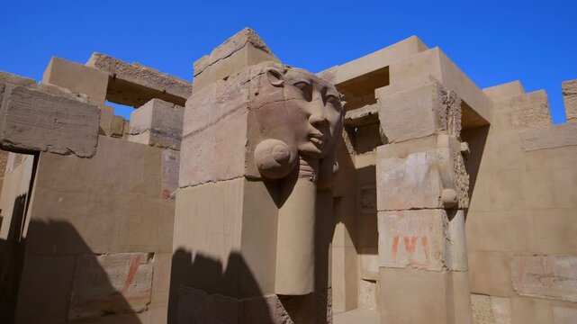 Close-up of ancient Egyptian Hathor-headed column capital with carved goddess face and cow ears on Elephantine Island temple ruins, Aswan, Egypt, against vivid blue sky.