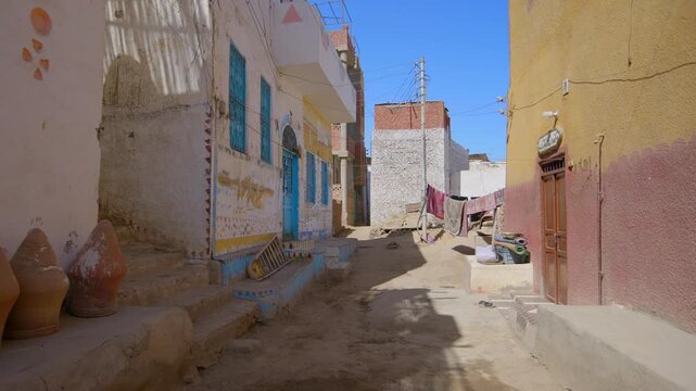 Colorful narrow alley in traditional Nubian village on Elephantine Island, Aswan, Egypt, with painted houses, blue windows, clay pots, laundry and sandy street under blue sky.
