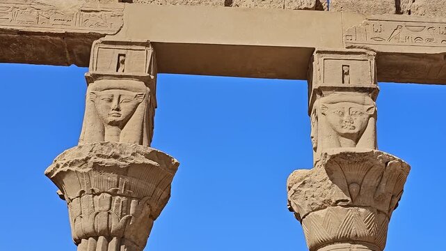 Magnificent colonnade of Philae temple with distinctive Hathor-faced capitals, Aswan, Egypt, Ptolemaic stone columns bearing carved faces under bright blue sky.