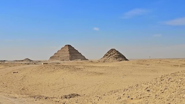 Panoramic desert view of Step Pyramid of Djoser and smaller ruined pyramid at Saqqara necropolis, Egypt, vast sandy landscape with ancient monuments under clear blue sky.
