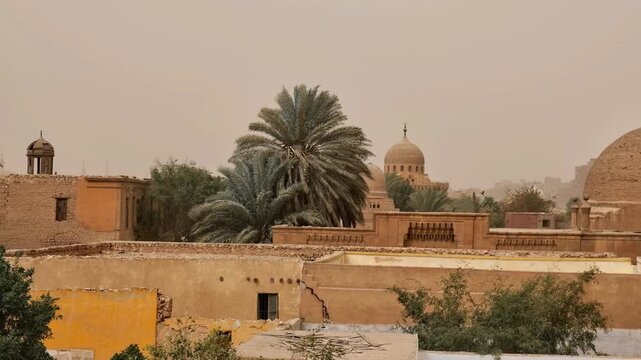 Medieval Islamic mausoleums and tomb structures of Cairo City of the Dead necropolis, Egypt, surrounded by palm trees and old buildings under orange dusty sky.