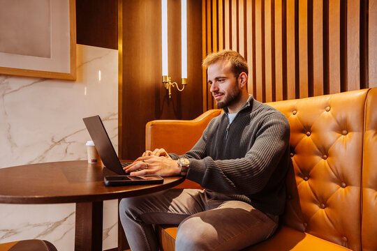 Young adult man smiling, working on laptop, sitting at table in stylish coffee shop.