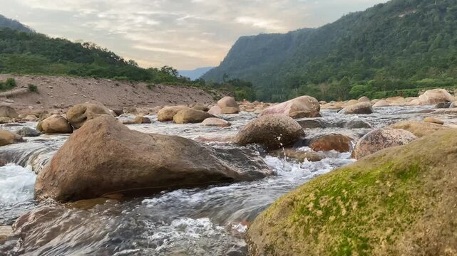 Peaceful river stream flows through boulders with mountain and sky - static shot.