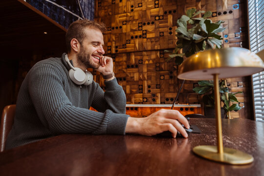 Young adult man laughing using computer mouse at wooden desk, enjoying remote work. Technology concept.