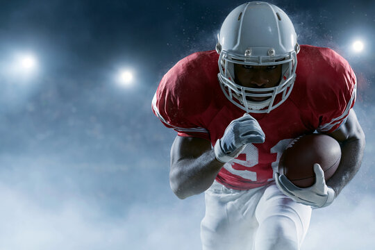 Athlete sprints forward, clutching football tightly. Red jersey, white helmet, focused intensity. Smoke and stadium lights enhance dramatic effect. Ideal for sports, energy, or action themes