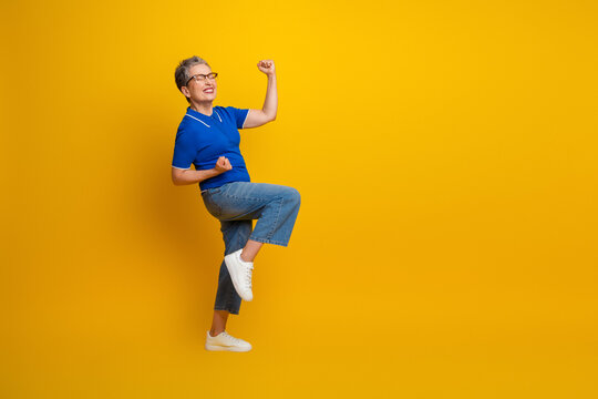 Energetic senior woman jumps in blue polo shirt against yellow background expressing joy and confidence