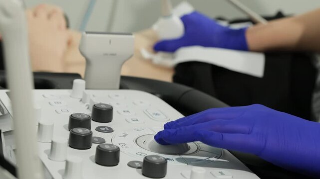 Gloved hand adjusts ultrasound console while patient lies during pelvic examination. Specialist fine tunes settings controling scan sequence