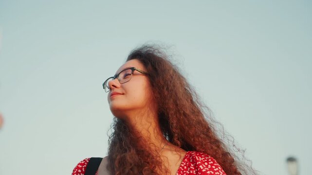 Closeup multiracial woman with glasses, curly hair against sunset skyline contemplative expression, windswept curls, red dress, soft golden light, rooftop towers in background, candid portrait