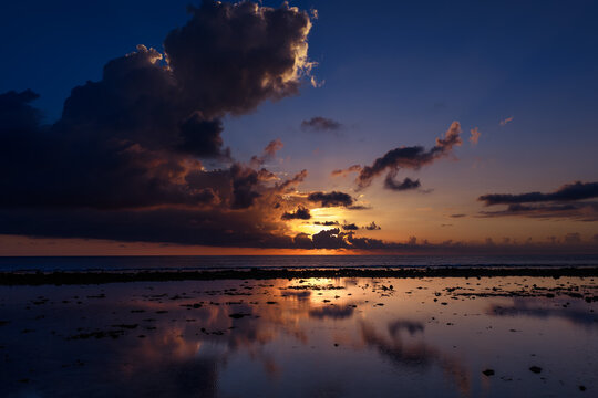 Aerial view of the sun setting behind dark clouds over the Indian Ocean at Mauritius Island with reflections on the water and rocky shore in Mauritius, Moka District, Mauritius.