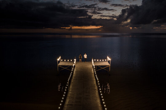 Aerial view of a wooden pier decorated with glowing string lights and two people standing at the end under a dark cloudy sky at dusk in Moka District, Mauritius.