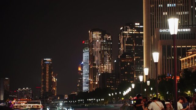 Night skyline waterfront promenade with lit streetlamps, highrise lights reflecting on river, evening crowd strolling, warm ambient glow, relaxed urban atmosphere, distant traffic and landmark