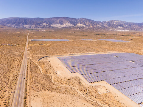 Aerial view of a large solar farm with rows of photovoltaic panels in the desert landscape near a straight road and mountains in Lucerne Valley, California, United States.