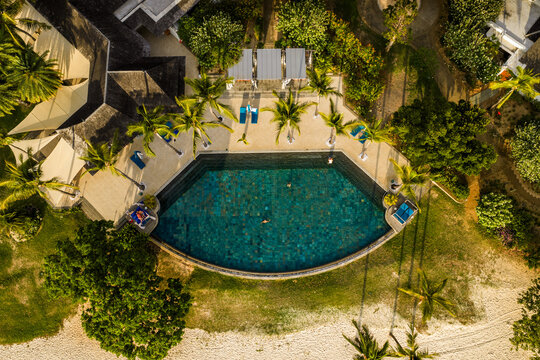 Aerial view of a semi-circular swimming pool with people swimming surrounded by palm trees and sun loungers at a luxury resort in Mauritius, Moka District, Mauritius.
