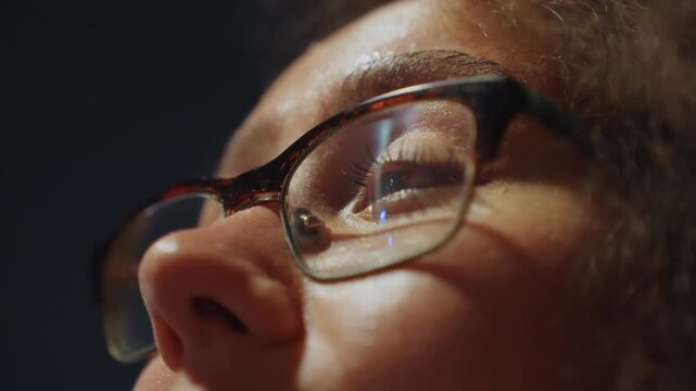 Student studying with reflective glasses. Face with glasses glowing softly in subdued lighting environment. Close view of student deep in thought with illuminated spectacles in lowlight room