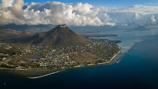 Aerial view of a coastal settlement at the base of a majestic mountain peak under a cloudy sky with turquoise ocean waters Mauritius, Moka District, Mauritius.
