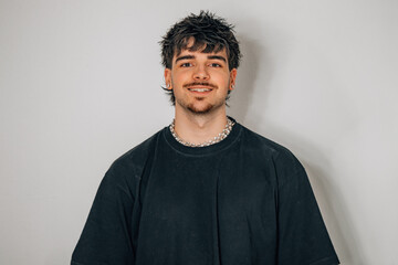 Obraz premium Young man with dark textured hair and a silver chain necklace smiling at the camera, wearing a black t-shirt against a gray studio background