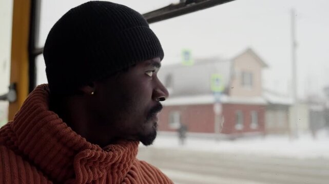 Black man gazing out bus window, snow falling over suburban street, garbage truck passing and houses blurred outside, wearing knit beanie and chunky orange sweater, contemplative expression, winter