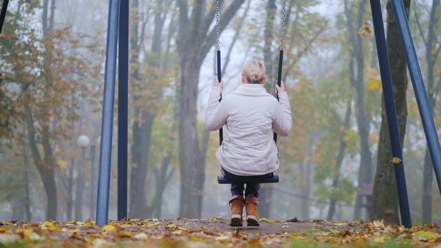 Woman sitting on swing in autumn park with fallen leaves, slow motion, peaceful mood.