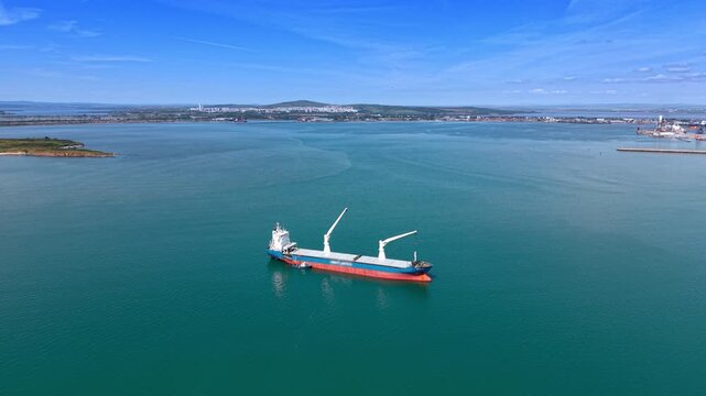 Approaching the ship equipped with two large heavy-lift cranes. Dry cargo vessel on the waterscape near the port. Aerial view.
