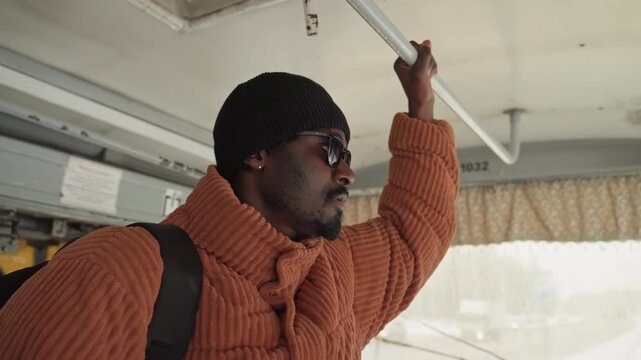 Black man standing on bus, wearing chunky orange sweater and beanie, gripping overhead rail as snowy city moves past the window backpack suggests student on morning commute, thoughtful expression,