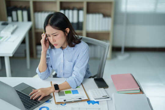 Stressed asian business woman experiencing a severe headache and burnout while working on her laptop in a modern office, overwhelmed by financial data and paperwork