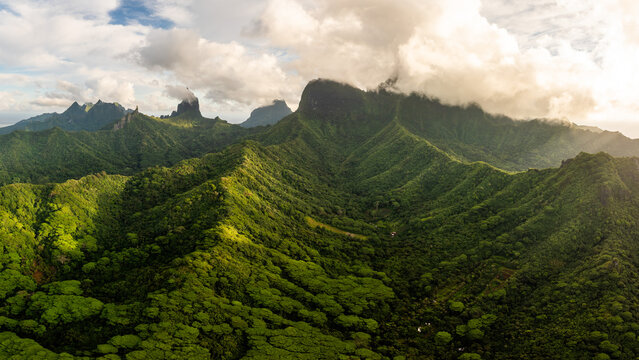 Aerial view of the lush volcanic peaks of Moorea and dense tropical forests under a cloudy sky in Moorea-Maiao, Windward Islands, French Polynesia.