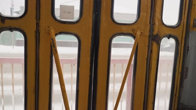 Yellow folding gate frames snowy street, wet metal step with circular studs and rust blurred pastel houses and parked car beyond window give quiet morning commuter mood, textured grime and salt
