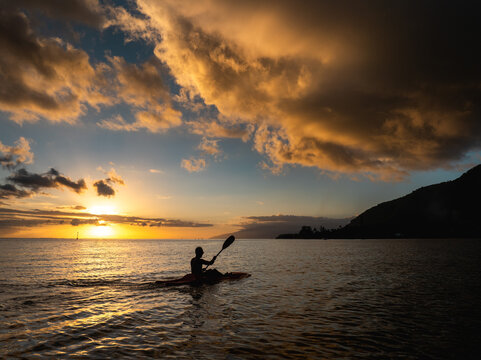 Aerial view of a person paddling a kayak on calm ocean waters during a golden sunset with dramatic clouds and the silhouette of Tahiti Moorea-Maiao, Windward Islands, French Polynesia.