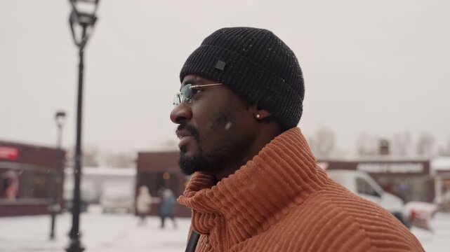 Black man profile in snowy square, closeup side view with glasses and knit beanie, snowflakes resting on beard and cap, orange truck and lampposts softly blurred in background, thoughtful gaze,