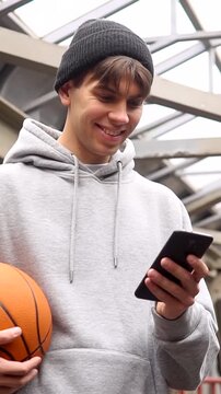 Young man basketball player with headphones holding ball using smartphone after training. Urban background. Vertical video.
