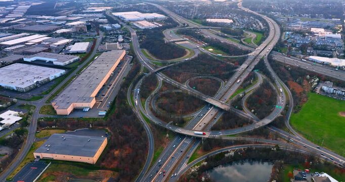 Outskirts of the modern city with warehouses and large interchange. Multiple cars move by the motorways and road loops. Aerial view.
