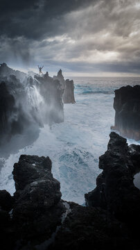Aerial view of a person standing on volcanic cliffs with arms raised as waves crash against the dark rocks under a dramatic sky in La Reunion, Nouvelle-Aquitaine, France.