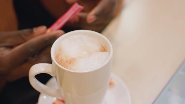 Black hands stirring frothy cappuccino on ceramic cup and saucer, closeup of hands in warm sweater adding sugar packet, soft morning light, intimate cafe vibe with slow motion stirring, aromatic steam