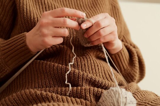 A person sits comfortably on a couch and knits using a ball of yarn and knitting needles. The activity takes place in a cozy indoor space during the afternoon