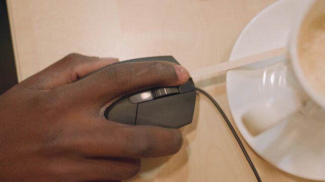 Overhead black hand on mouse with straw across buttons, designer testing click sensitivity beside latte and saucer, inventive studio moment blending creativity and casual coffee break