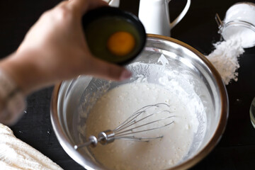 Fototapeta premium Pancake batter preparation on a dark background.Whisking liquid batter in a metal bowl on a dark background, raw eggs nearby. Cooking process concept.