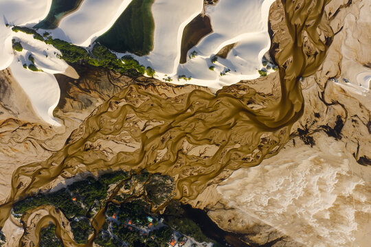 Aerial view of Parque Nacional dos Lencois Maranhenses with a winding brown river flowing through white sand dunes and green vegetation in Lencois Maranhenses, MA, Brazil.