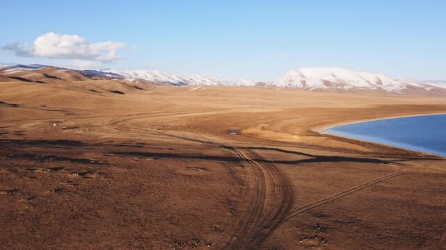 Vast aerial perspective of the high-altitude steppe and dirt tracks along the shores of Song-Kul Lake, Kyrgyzstan. Snow-capped mountains frame the remote Central Asian nomadic landscape