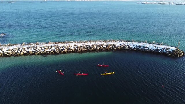aerial drone view of colorful kayaks paddling in clear turquoise arctic waters next to a rocky harbor in Lofoten Islands, Norway. Active outdoor adventure in the Scandinavian fjords