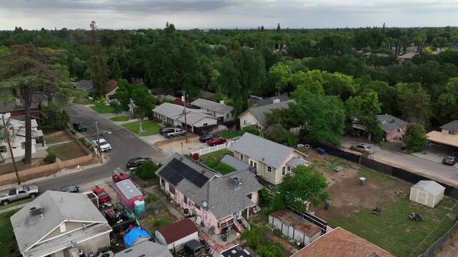 Residential neighborhood in Modesto California with suburban houses with solar panels and lush green trees along quiet street during cloudy day in Central Valley, USA. Aerial rising shot.